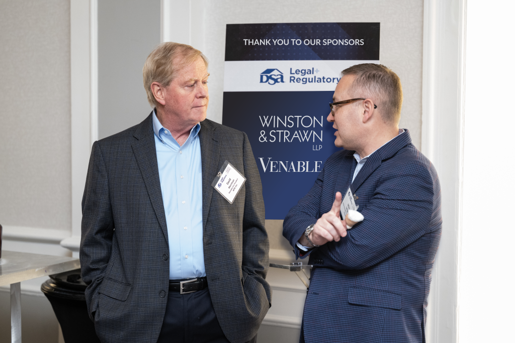 two men speaking to each other in front of a poster with sponsorship logos on it
