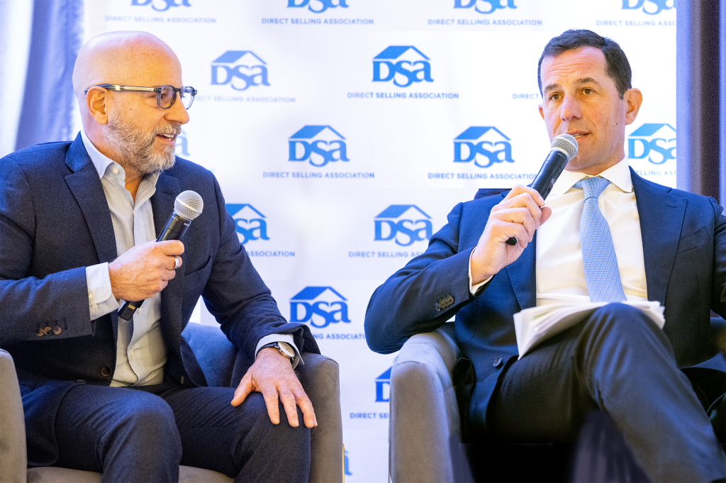 Two men sitting in chairs speaking at a DSA event while holding microphones. A DSA step and repeat in the background.